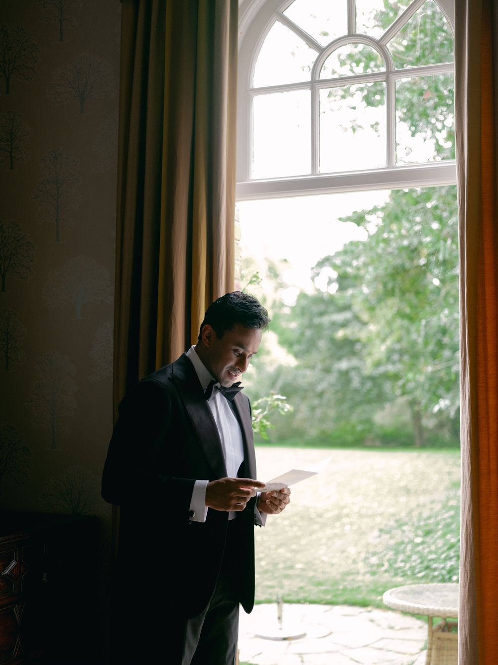 Groom in black tuxedo reading a letter by a large arched window with garden views on wedding day.