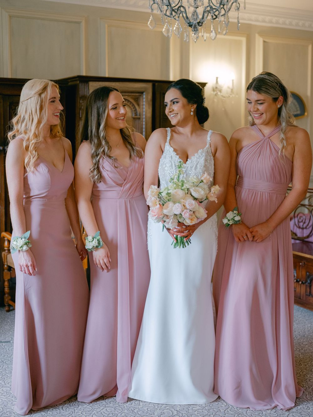 Bride in white lace gown with three bridesmaids in dusty rose satin dresses holding bouquet.