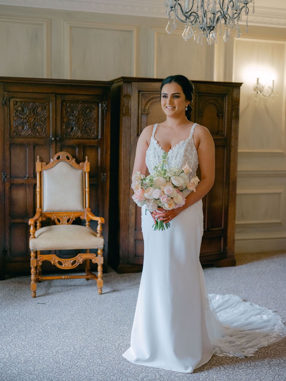 Bride in white lace mermaid wedding gown holding pink bouquet in elegant wood-paneled room.
