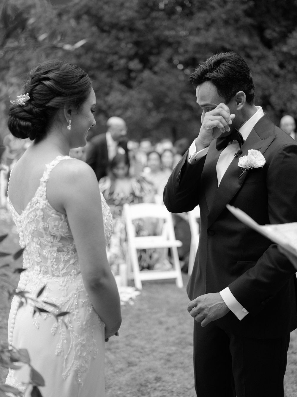 Emotional black and white wedding ceremony photo of groom wiping tears as bride faces him.
