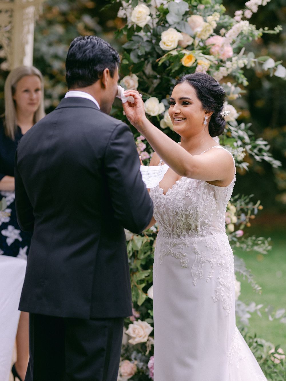 Bride smiling while placing wedding ring on grooms finger during outdoor ceremony.