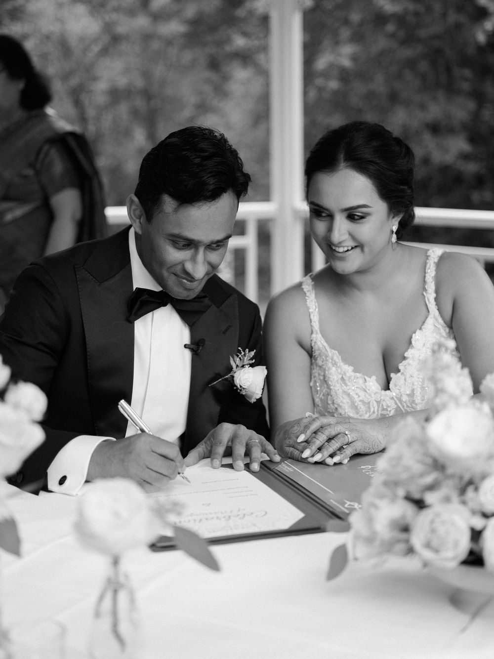 Bride and groom signing wedding certificate at reception table in black and white.