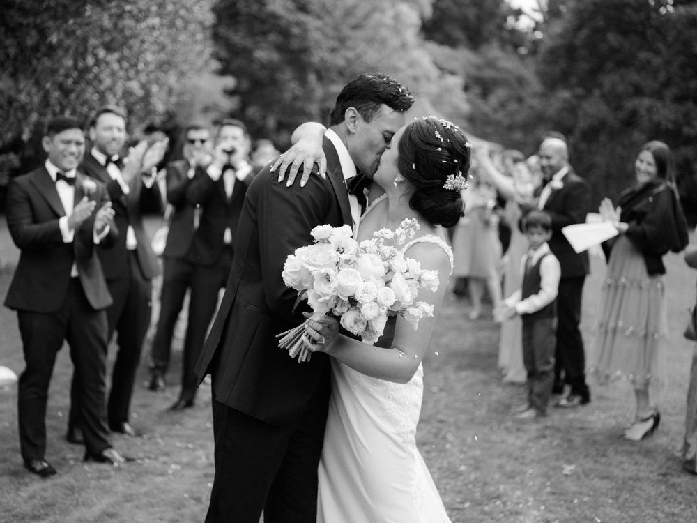 Black and white photo of newlyweds kissing while guests applaud outdoors at elegant wedding ceremony.