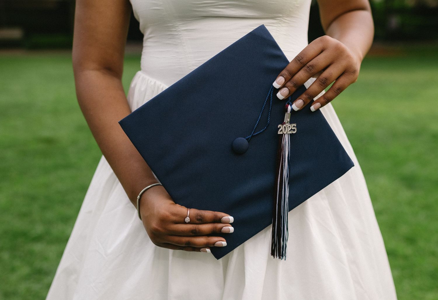 Navy blue clutch purse with tassel detail shown against a white dress and green outdoor background.