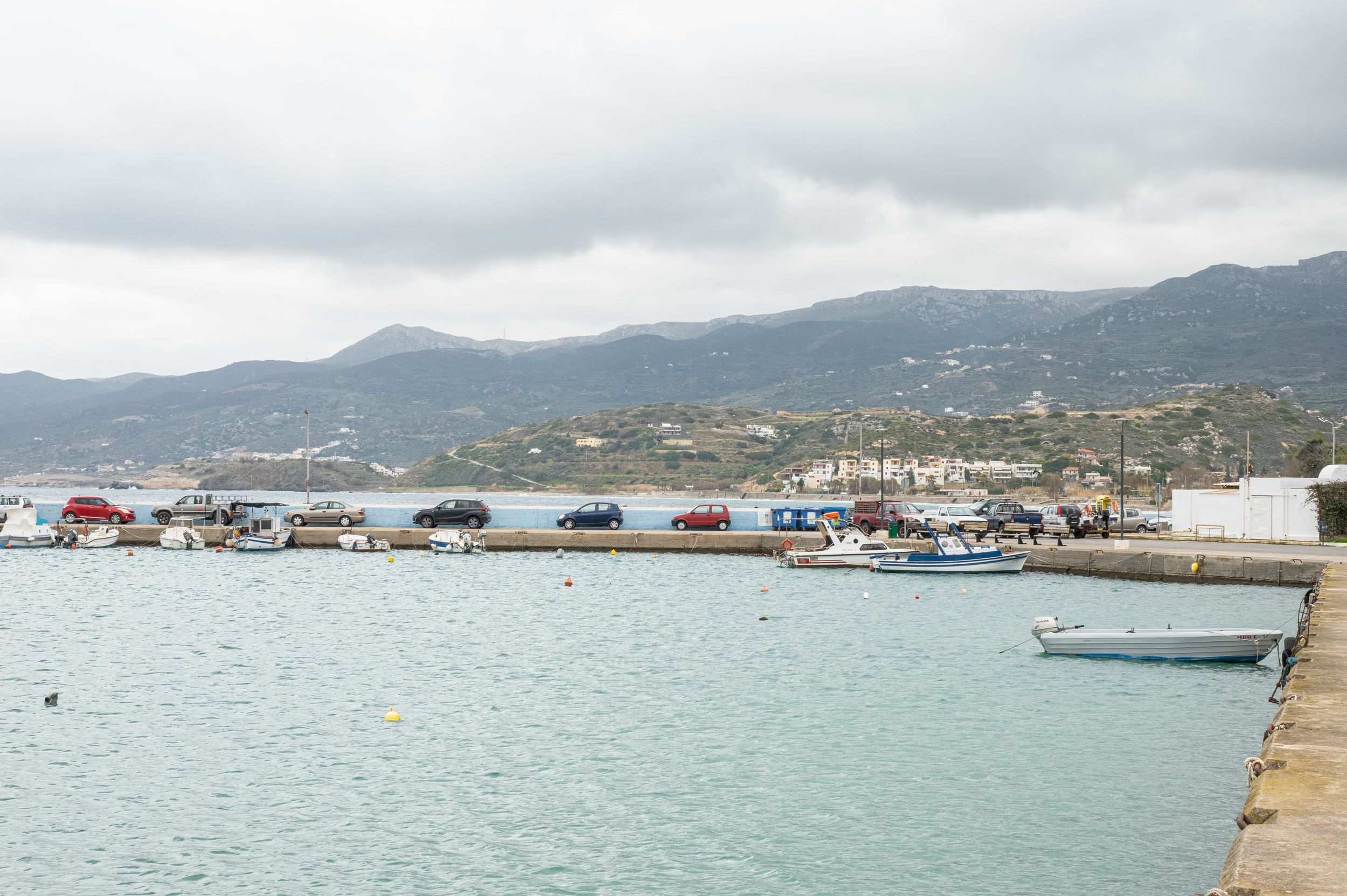 Coastal harbor view with fishing boats docked along turquoise waters against mountainous backdrop in cloudy weather.