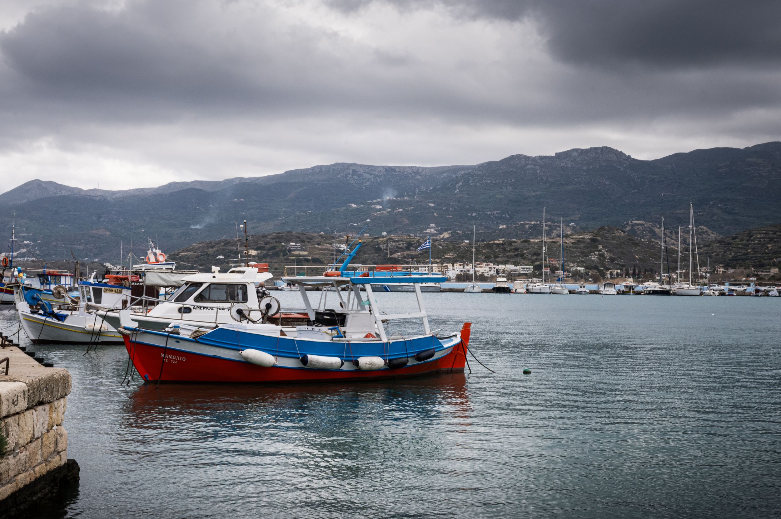 Fishing boats anchored in a harbor with mountains and cloudy skies in the background.