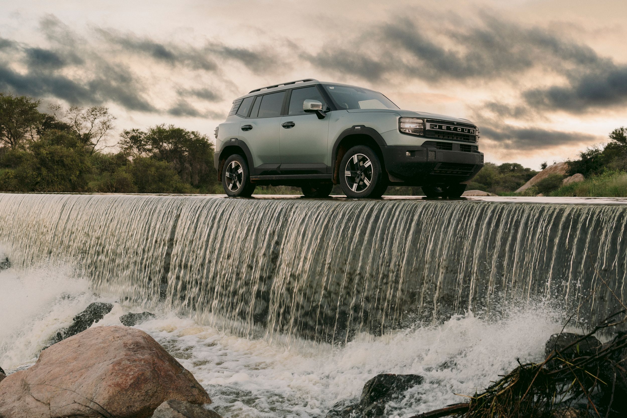 Silver Ford Bronco Sport SUV parked on a waterfall dam edge with dramatic cloudy sky at sunset.