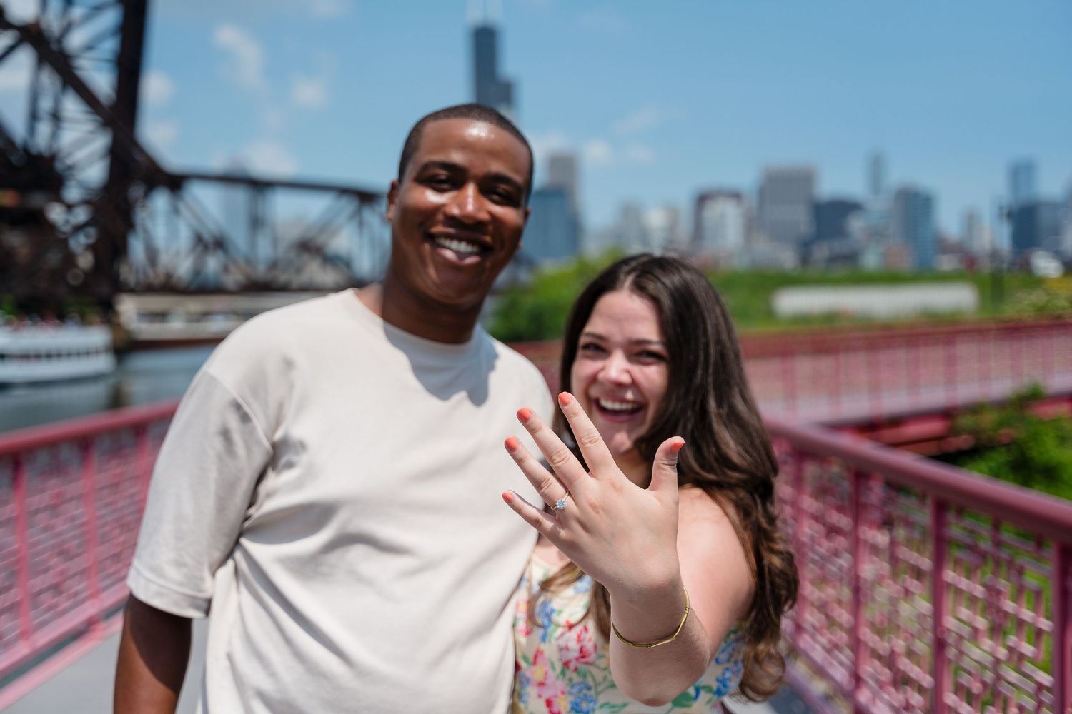 A happy couple stands on a bridge showing off an engagement ring with a city skyline visible in the background.