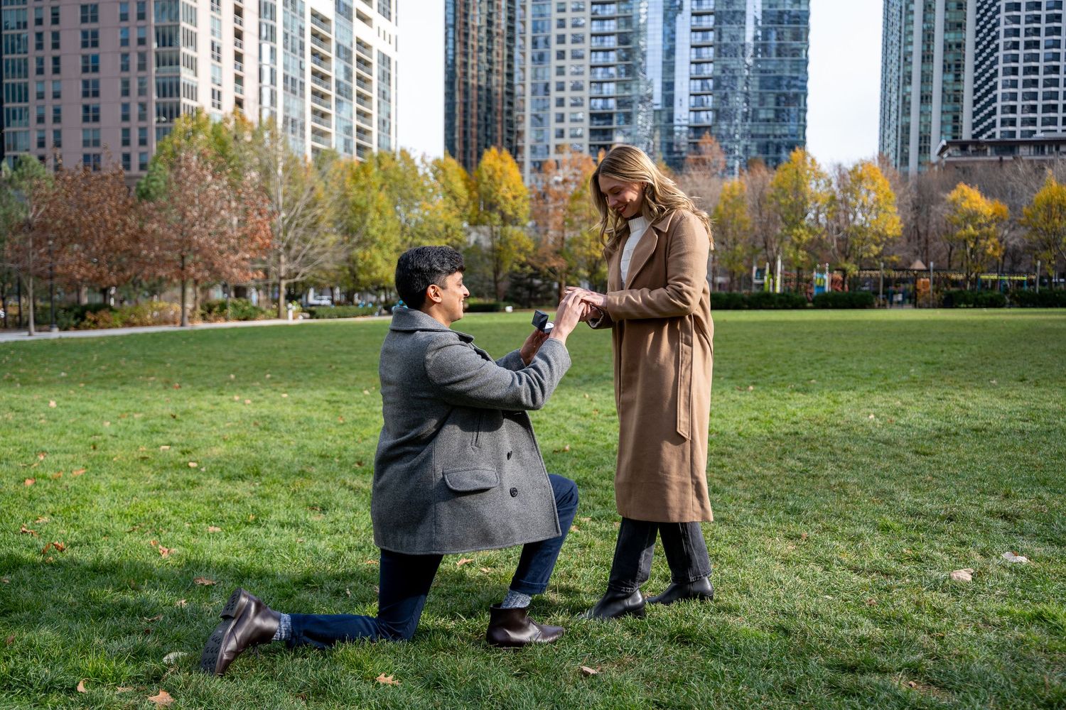 People relax on a grassy lawn in a city park surrounded by tall buildings on an autumn afternoon.