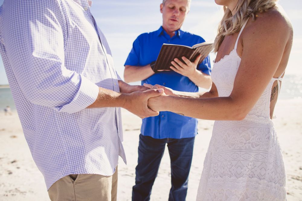 A couple exchanges rings during a beach wedding ceremony with an officiant in a blue shirt standing nearby.