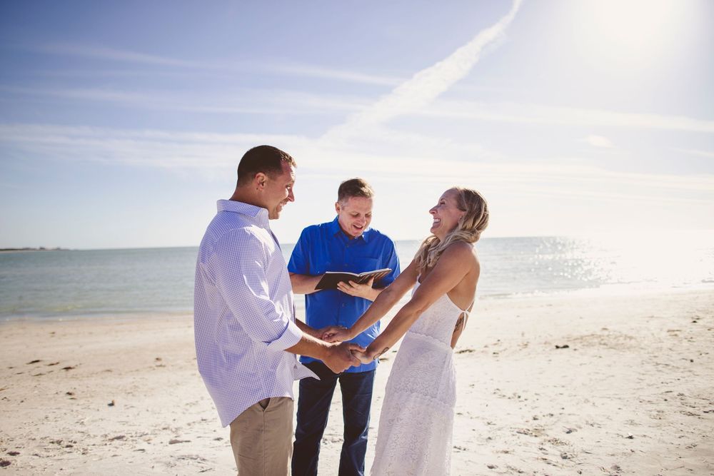 A couple participates in a beach wedding ceremony on a sunny day with white sand and ocean views.