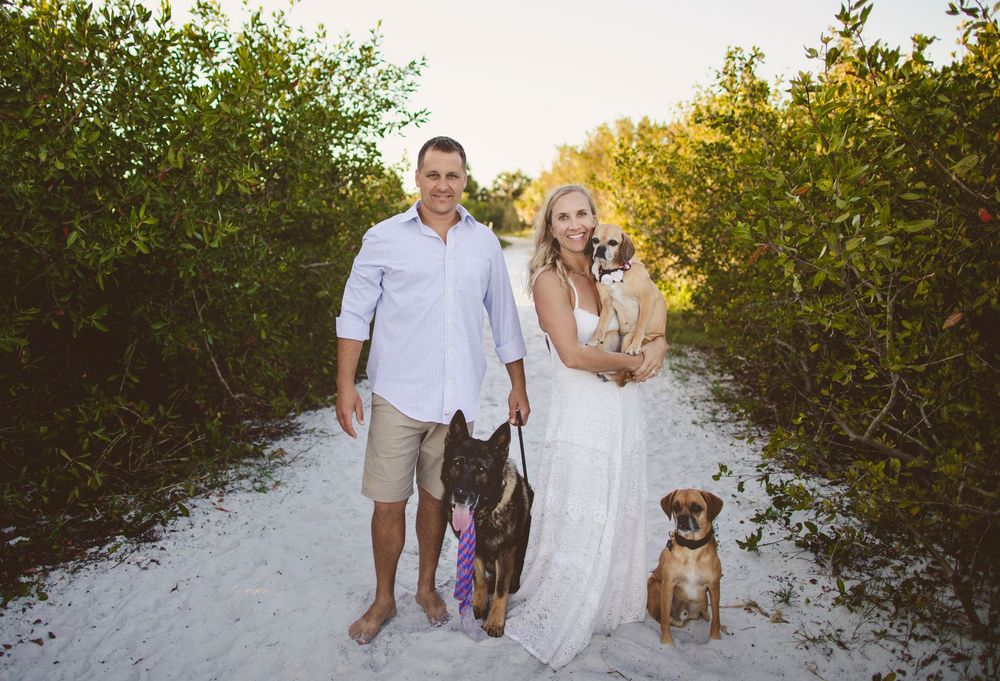 A couple stands on a sandy beach path with two dogs during golden hour sunset.