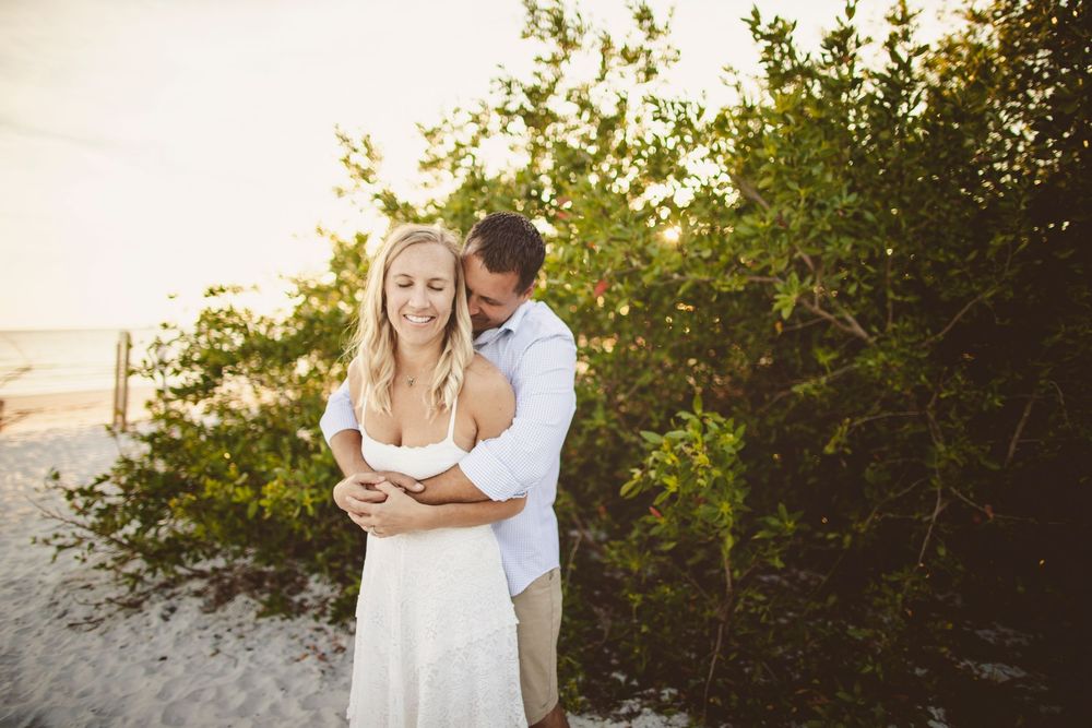 A couple embraces during a romantic sunset photoshoot on the beach with golden light filtering through trees.