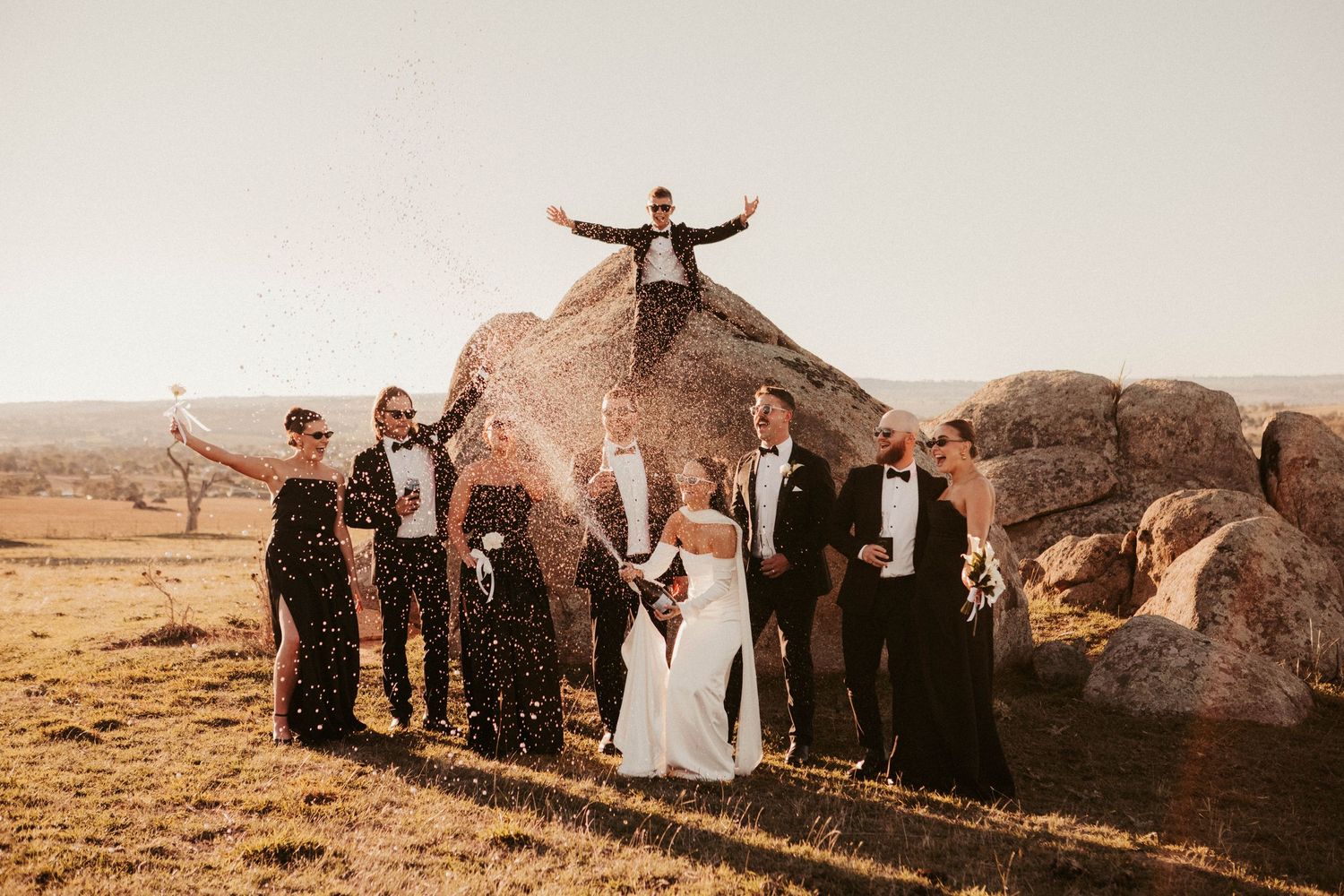 Wedding party celebrating outdoors on rocky terrain, bride and groom with guests in formal attire, man posing atop boulder.
