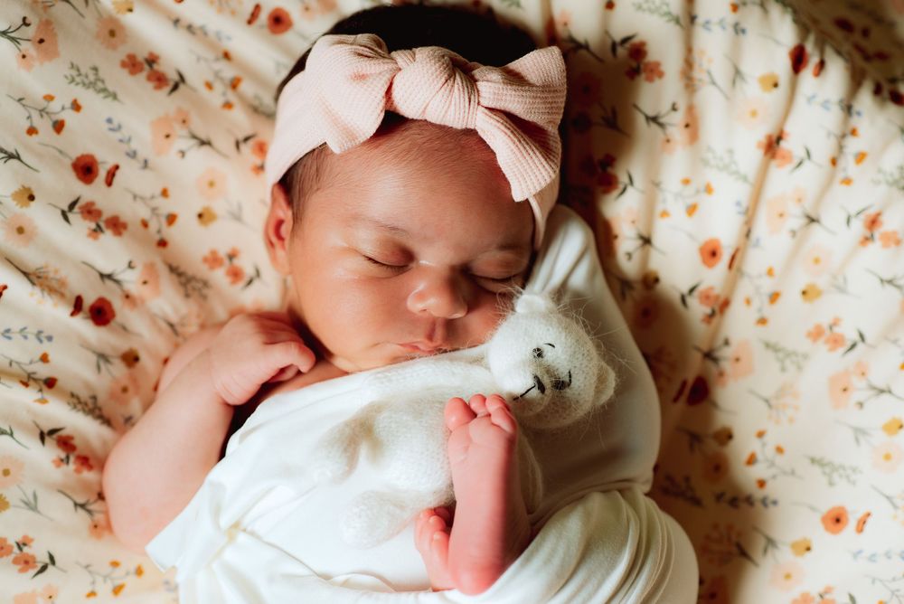 Newborn girl wrapped with tiny teddy bear and foot sticking out. Has a flower blanket backdrop