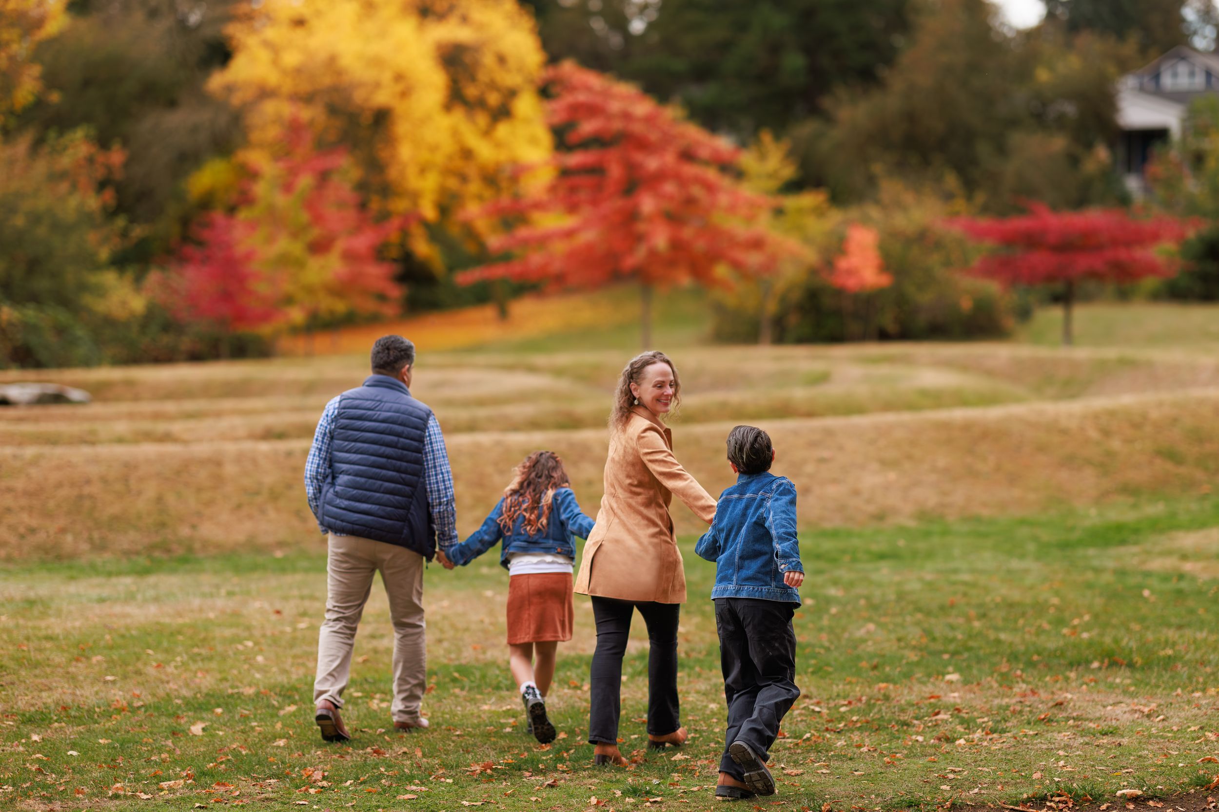 Family of four walking hand-in-hand through a colorful autumn park with vibrant red and yellow foliage.