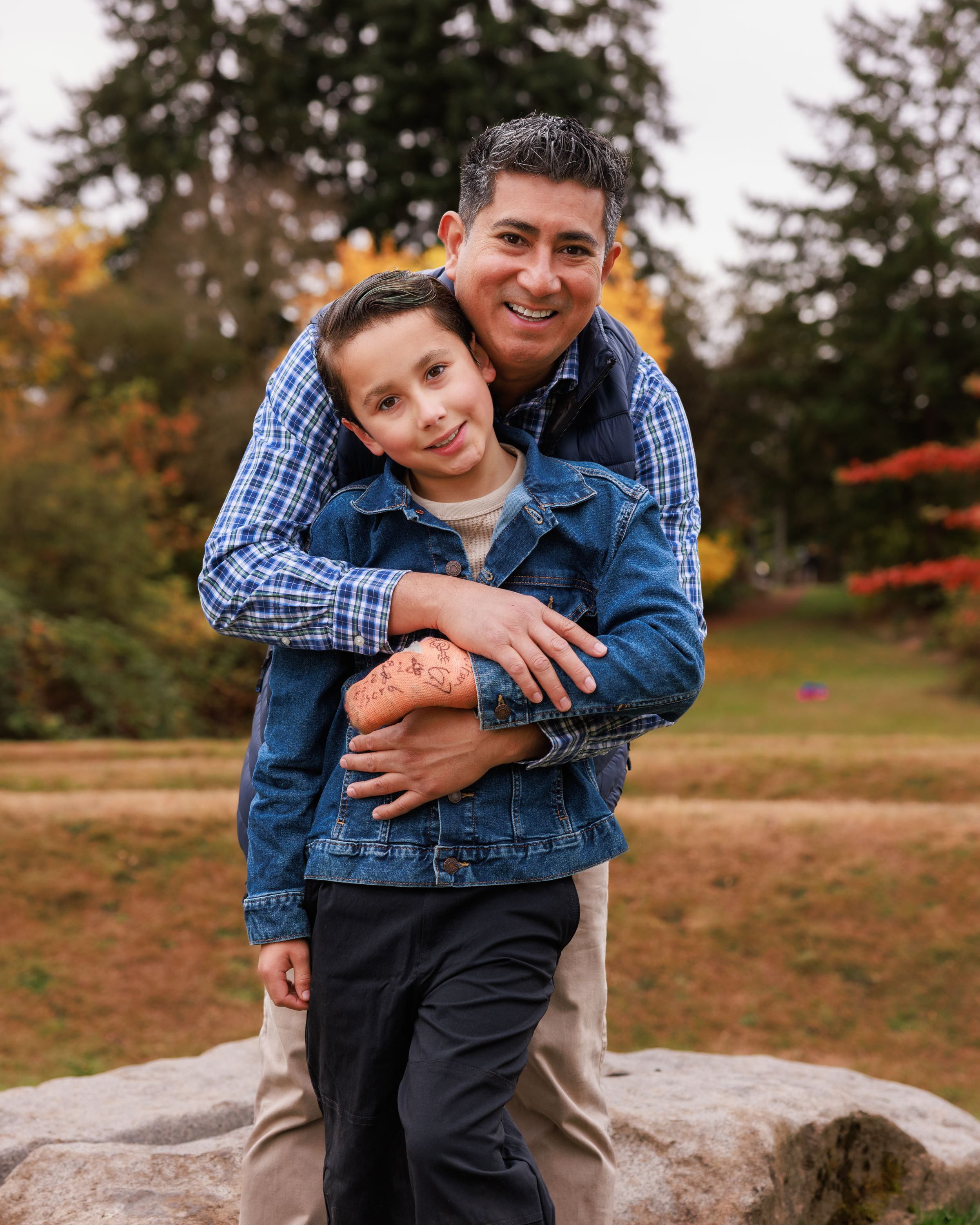 A father and son smile together in a park with autumn foliage, the man hugging the boy from behind on a rock.