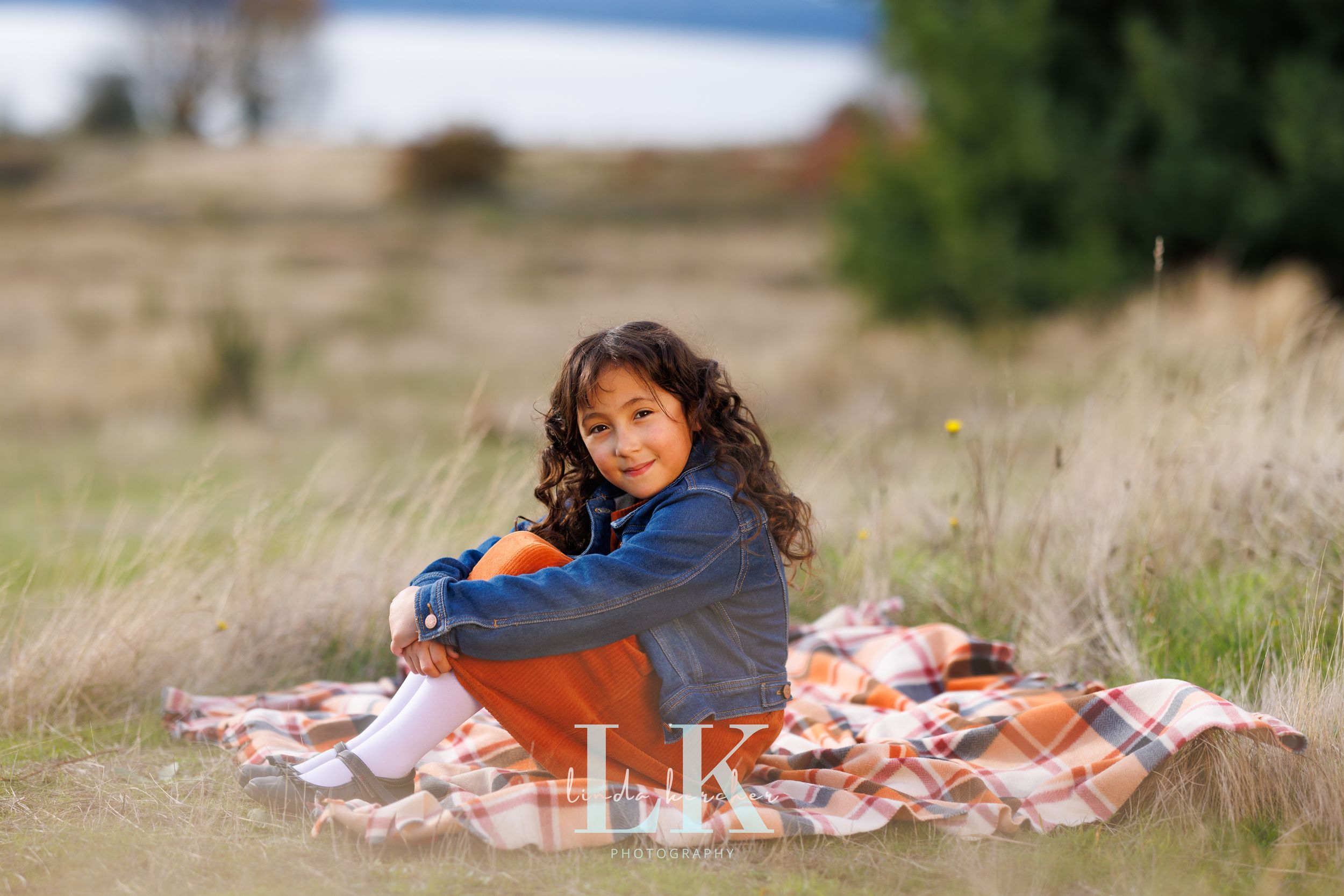A young girl sits smiling on a plaid blanket in a grassy field, wearing a denim jacket and orange pants.