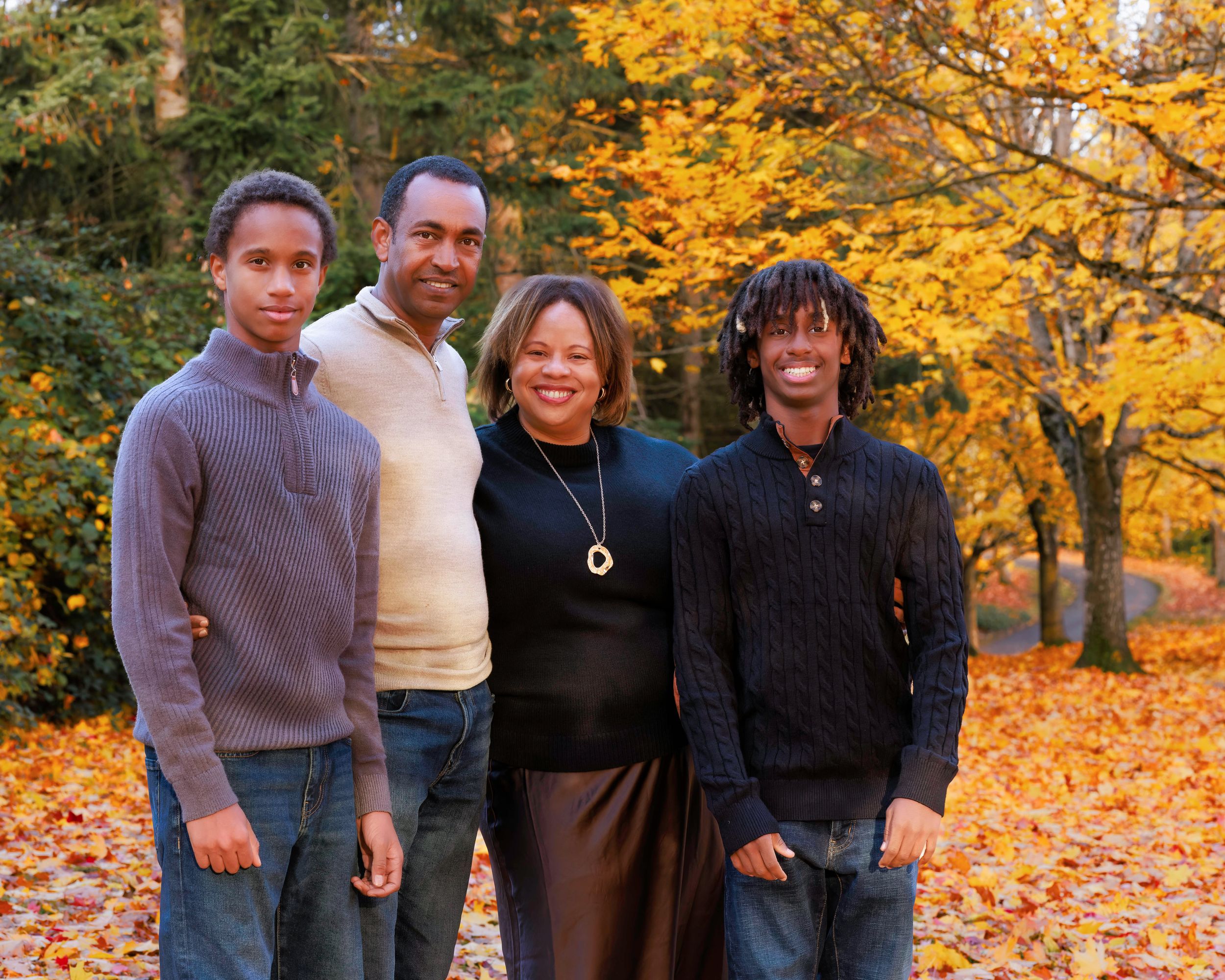 Family of four smiling together outdoors among colorful autumn foliage and fallen leaves in a park setting.