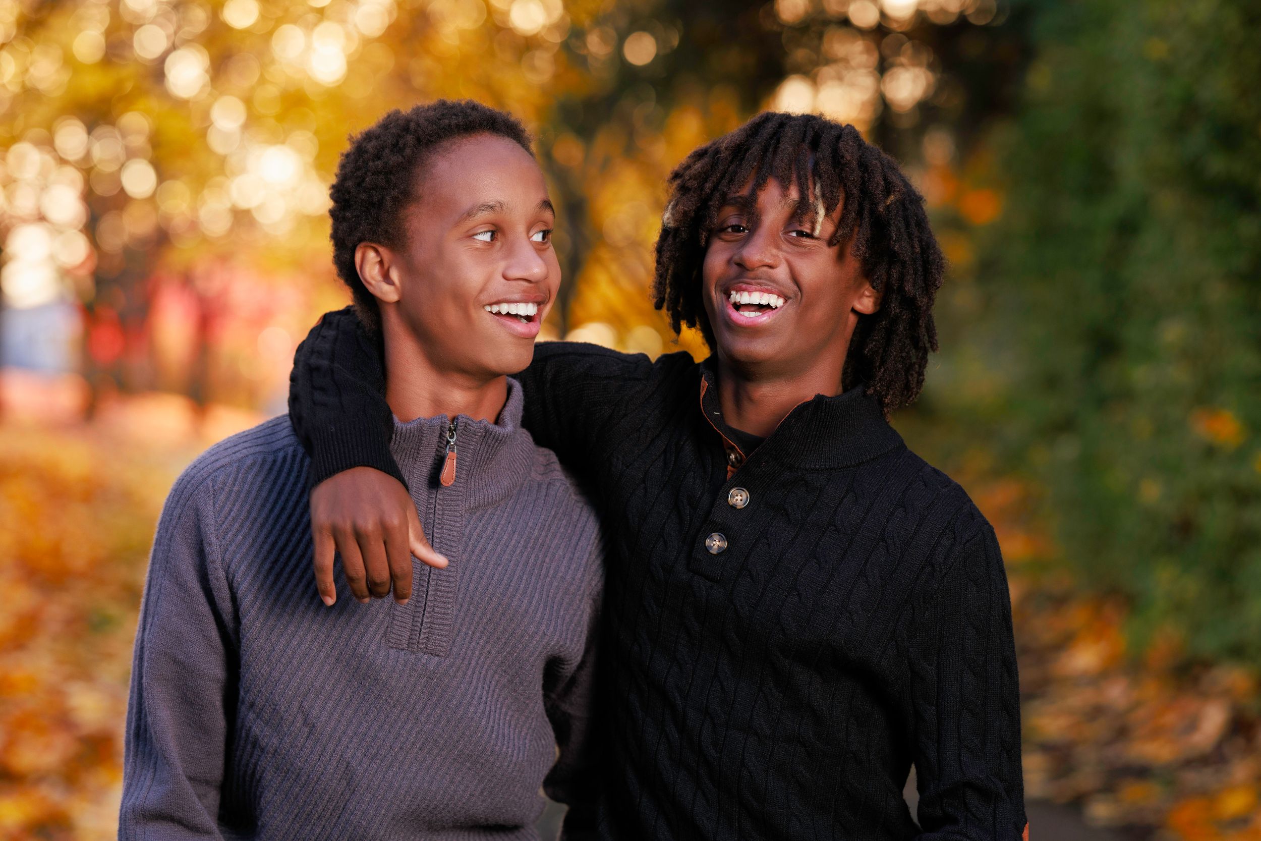 Two young Black men smiling and embracing outdoors among colorful autumn foliage.