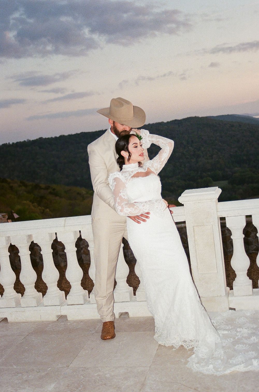 Bride and groom sharing romantic dip pose on white stone terrace with mountain and cloudy sky backdrop.