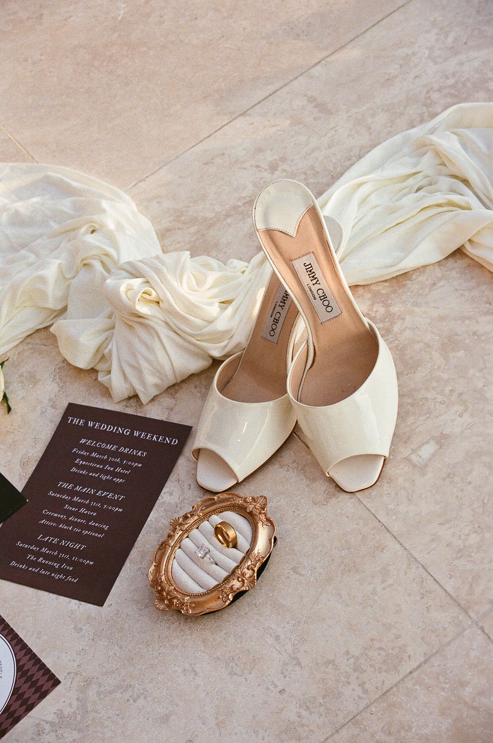 Jimmy Choo bridal heels with wedding rings in ornate gold dish beside wedding weekend itinerary card and white dress.