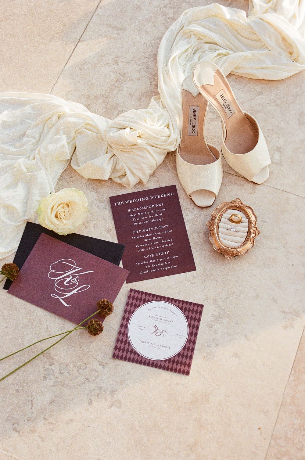 Flat lay of wedding stationery suite with ivory Jimmy Choo heels, roses, and silk fabric on tile.