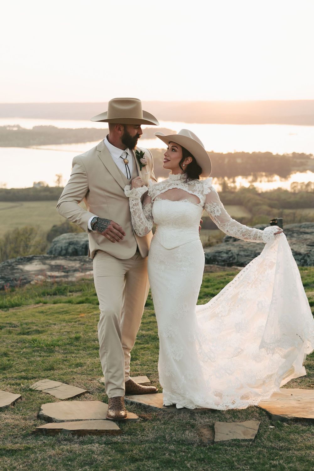 Western wedding couple in cowboy hats dance on hilltop, bride in lace gown holding train at golden sunset.