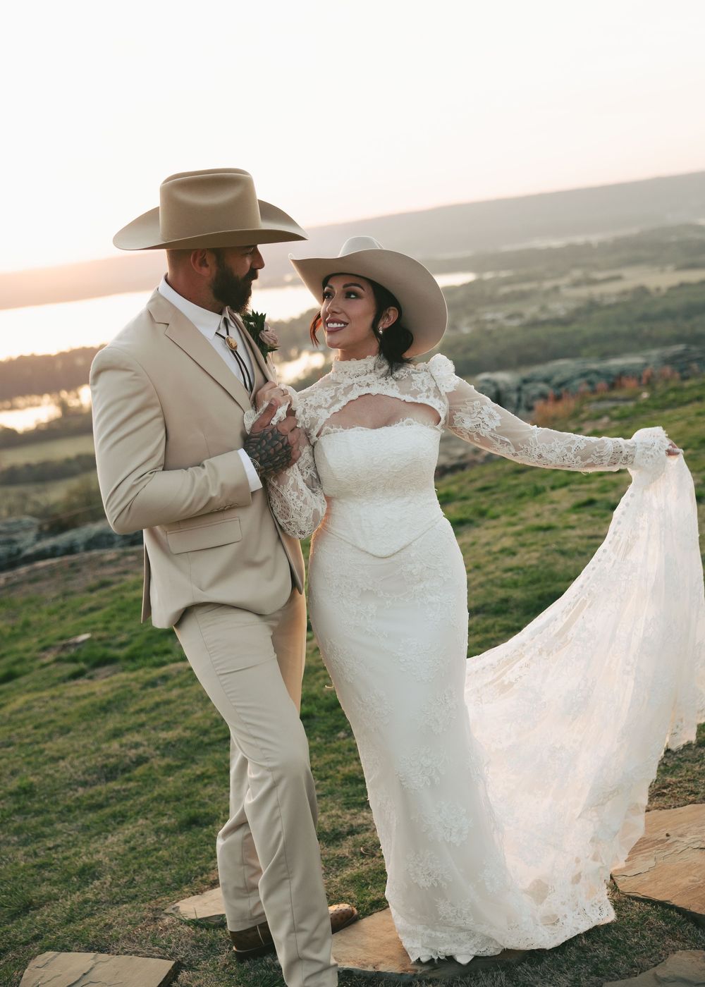 Smiling western wedding couple in cowboy hats pose at sunset, bride in long-sleeve lace gown holding flowing train.