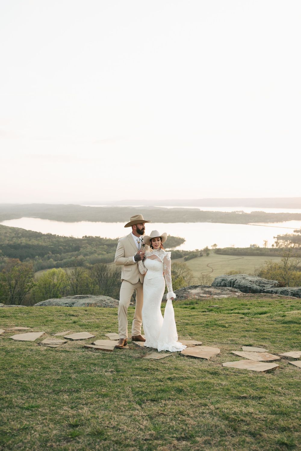 Western wedding couple in wide-brim cowboy hats stand together on hilltop overlooking scenic lake at dusk.