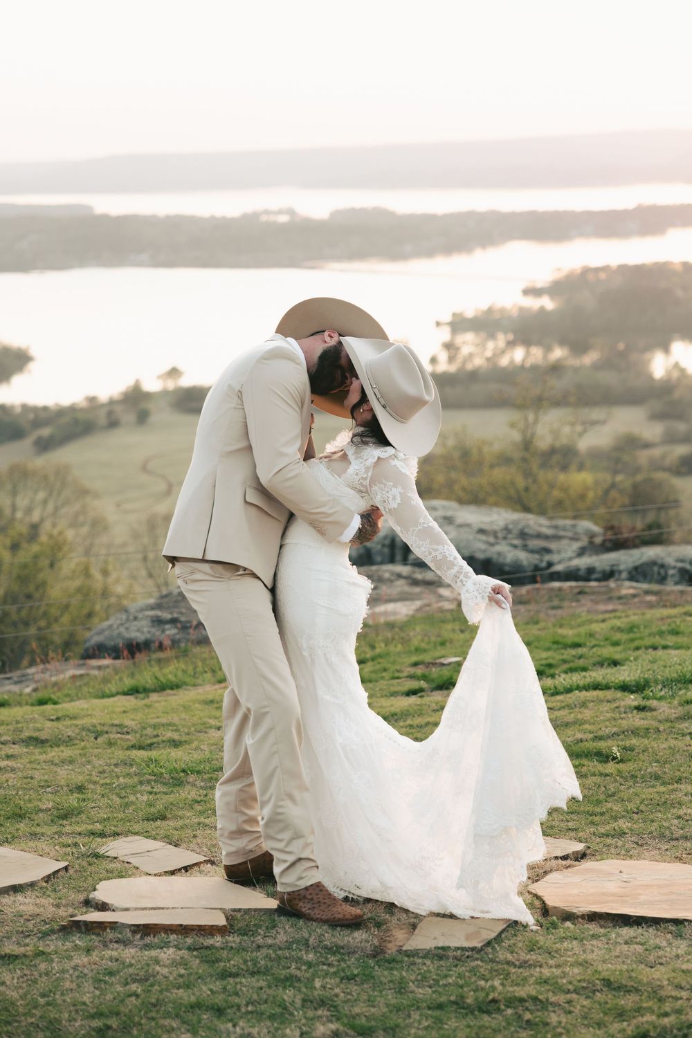 Western couple sharing passionate kiss on hilltop at sunset, bride in lace gown and groom in tan suit with cowboy hats.