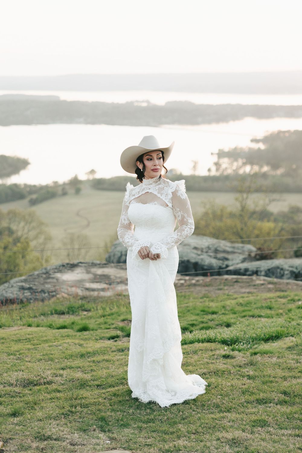 Bride in white lace gown and cowboy hat stands on grassy hilltop with scenic lake view at sunset.