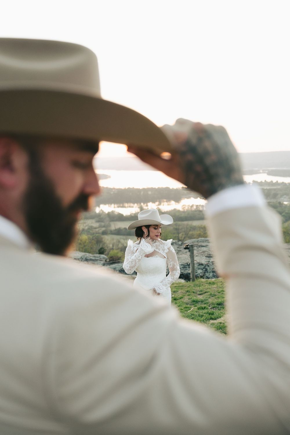 Groom in white suit adjusts cowboy hat while bride in white lace dress stands in background outdoors.