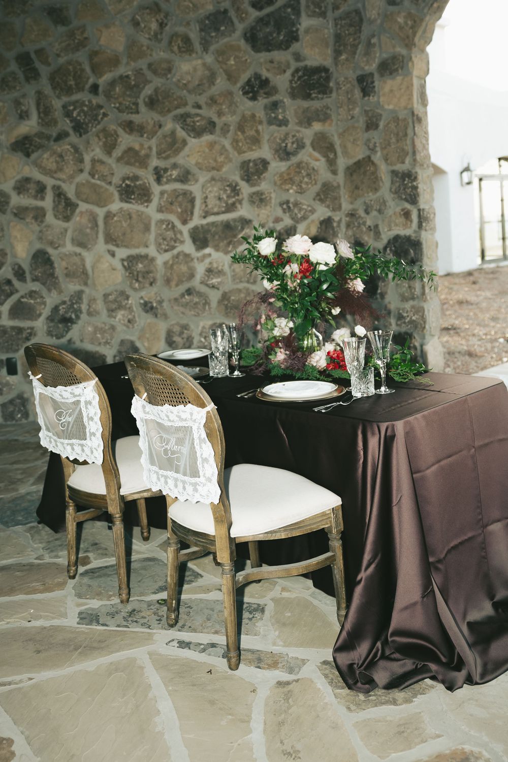 Elegant sweetheart table with dark linen, floral centerpiece, and cane-back chairs against rustic stone wall.