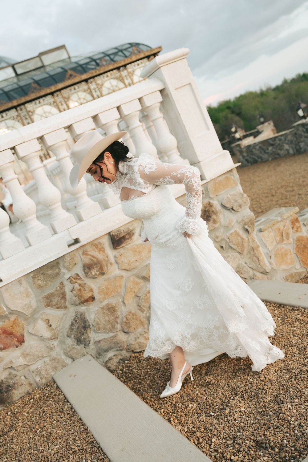 Bride in white lace wedding gown and hat poses beside stone balustrade at rustic outdoor venue.