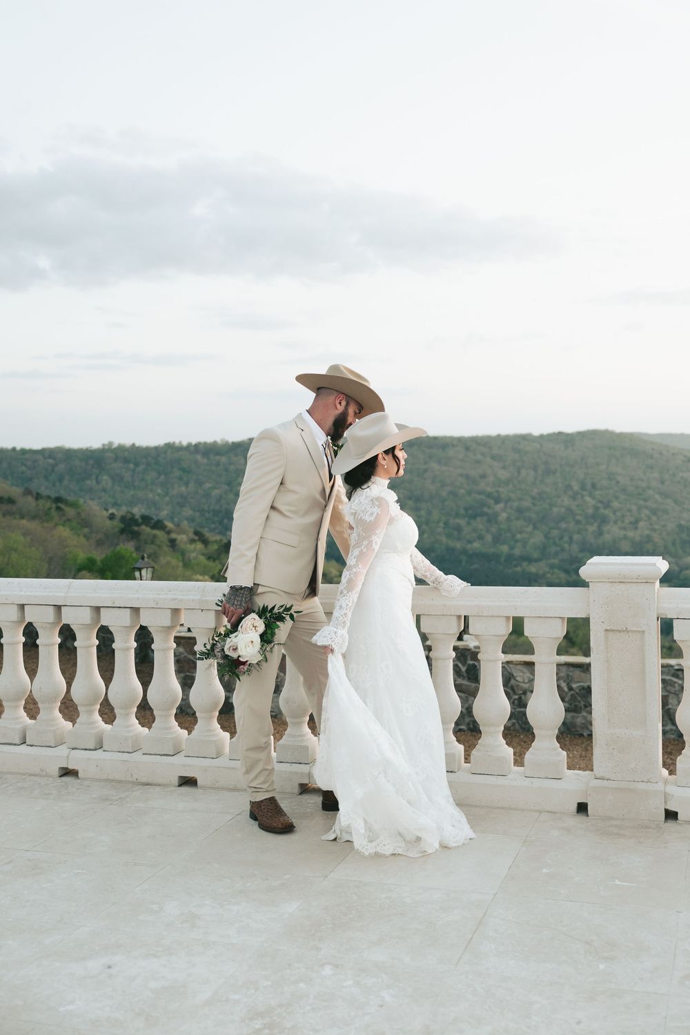 Bride and groom in matching cowboy hats walk along white stone balcony terrace with mountain views.