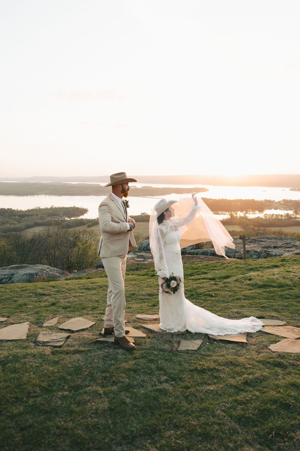 Western bride in lace dress and cowboy hat lifts veil in wind beside groom on scenic hilltop at sunset.