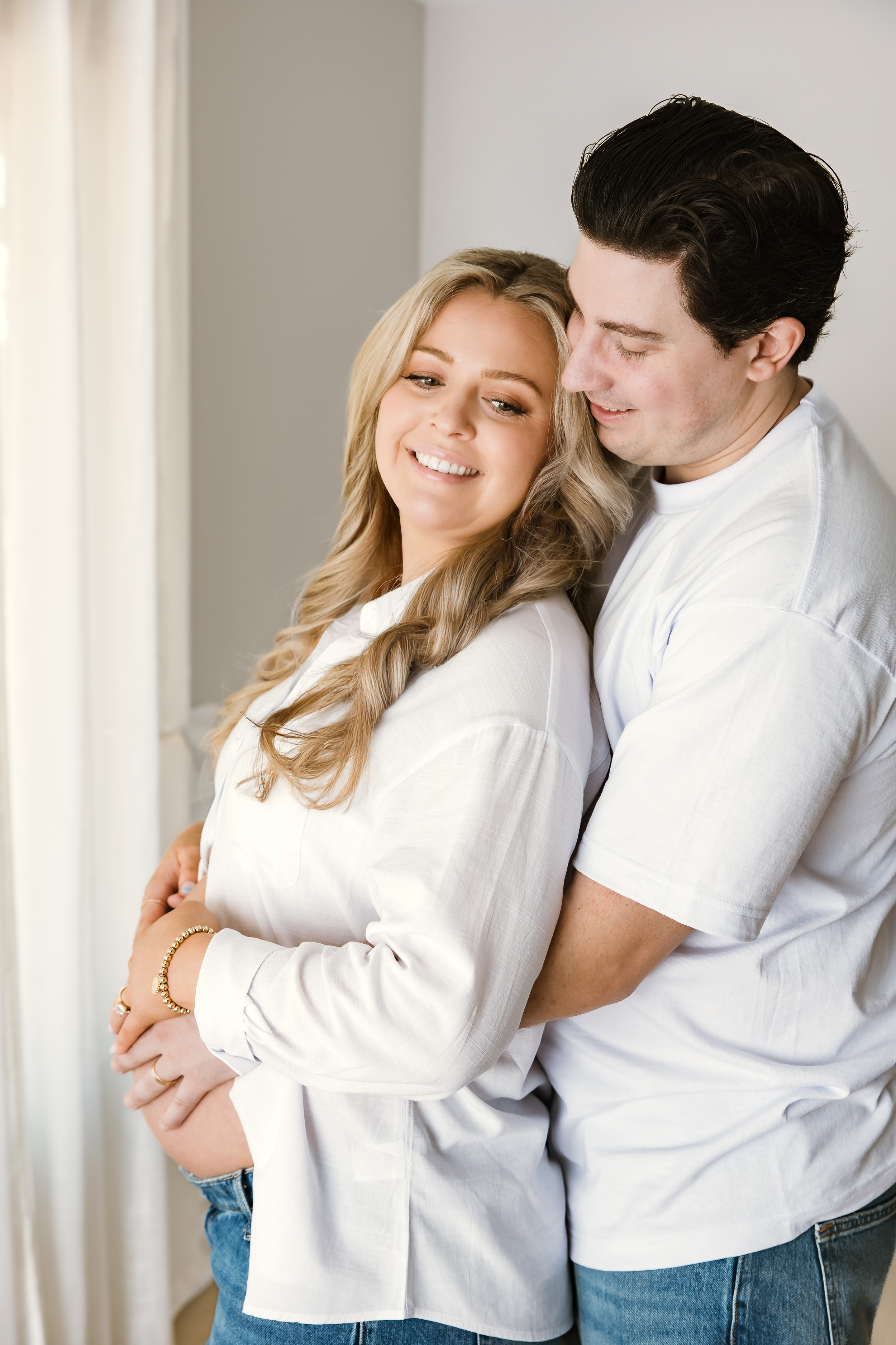 Smiling pregnant woman in white shirt embraced from behind by partner near bright window.