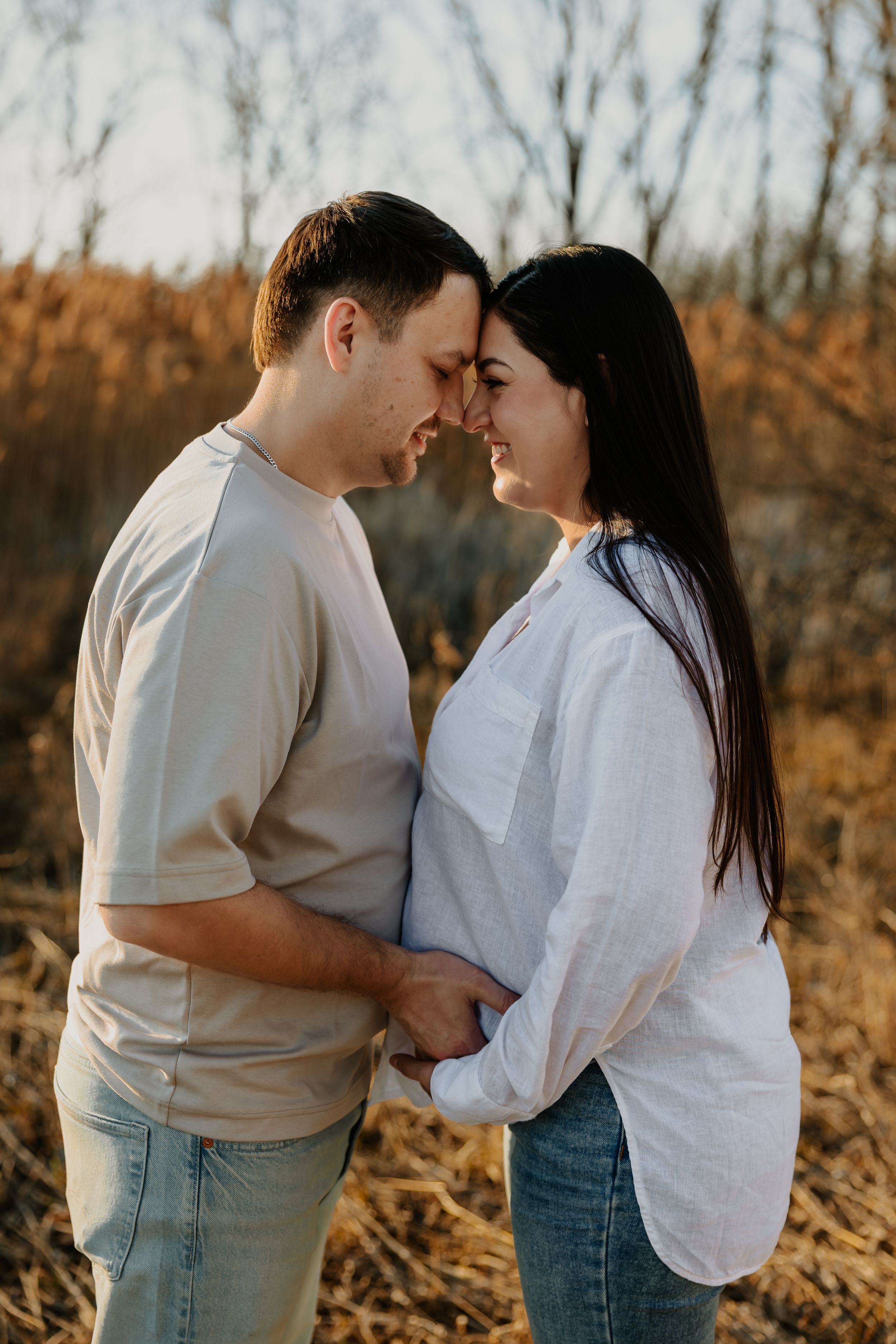 Loving couple touching foreheads tenderly while holding hands in a golden dried grass field at sunset.