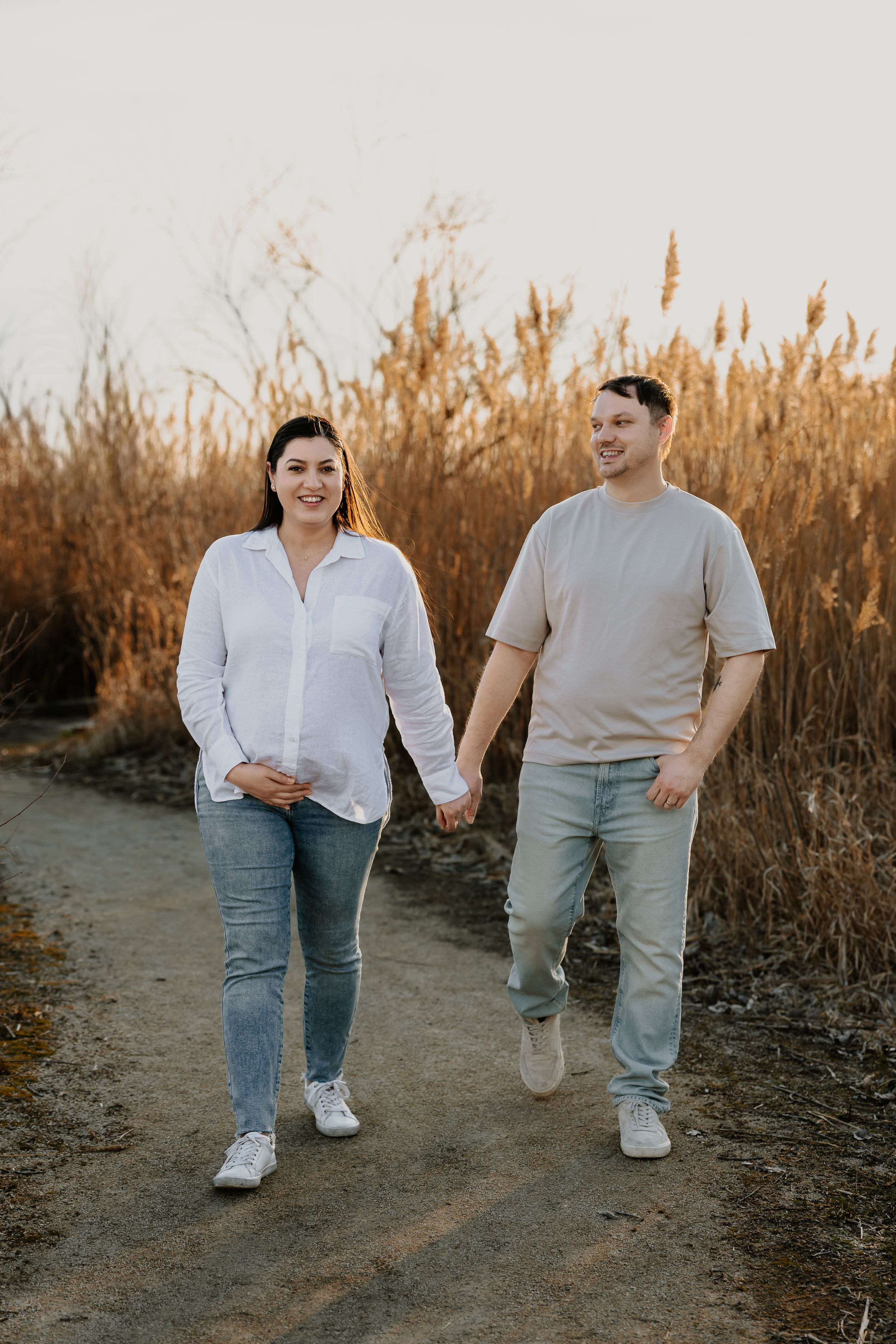 Pregnant couple walking hand in hand along a path through tall golden grass in warm sunset light.