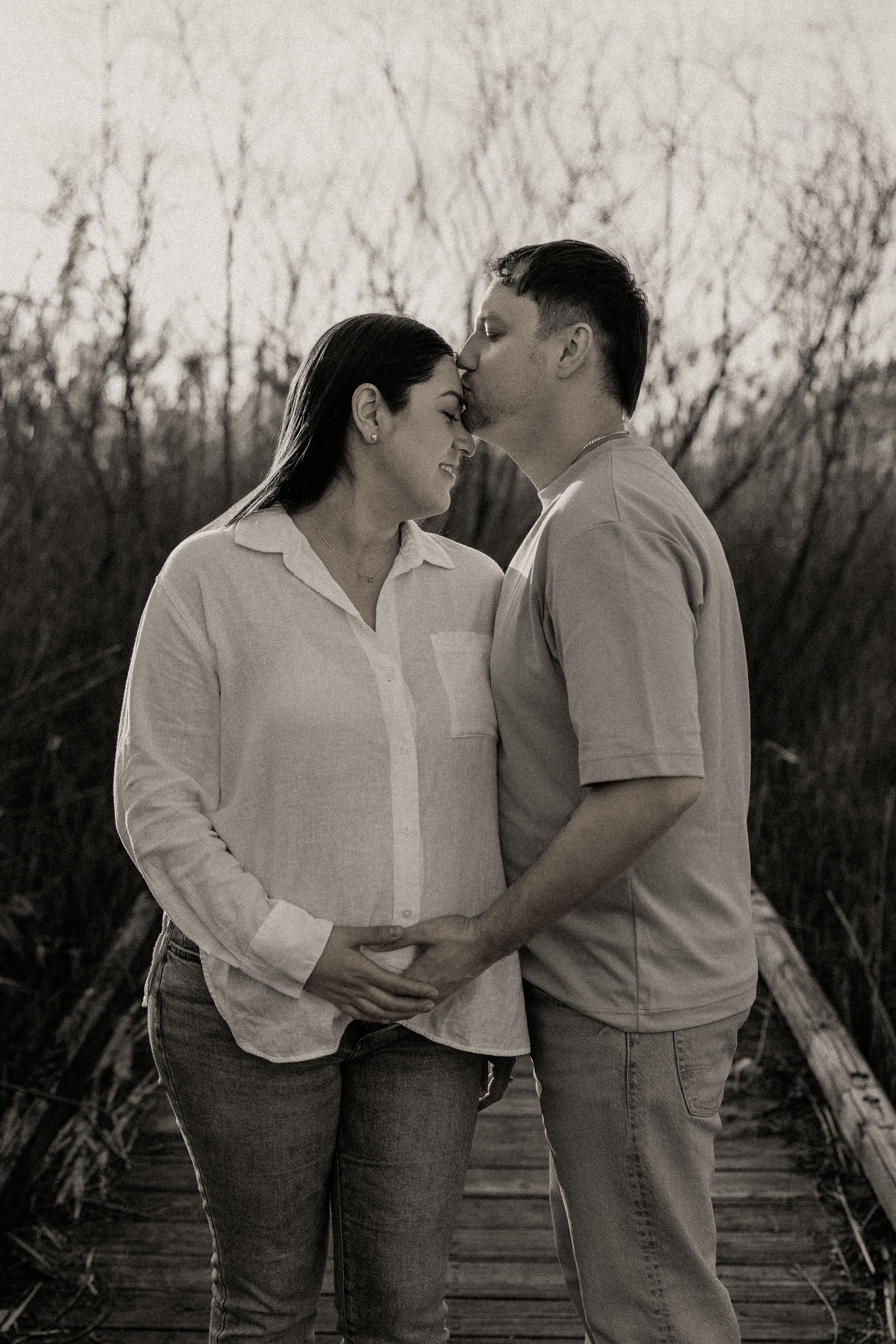 Black and white photo of expecting couple embracing on a wooden bridge surrounded by tall winter grass.