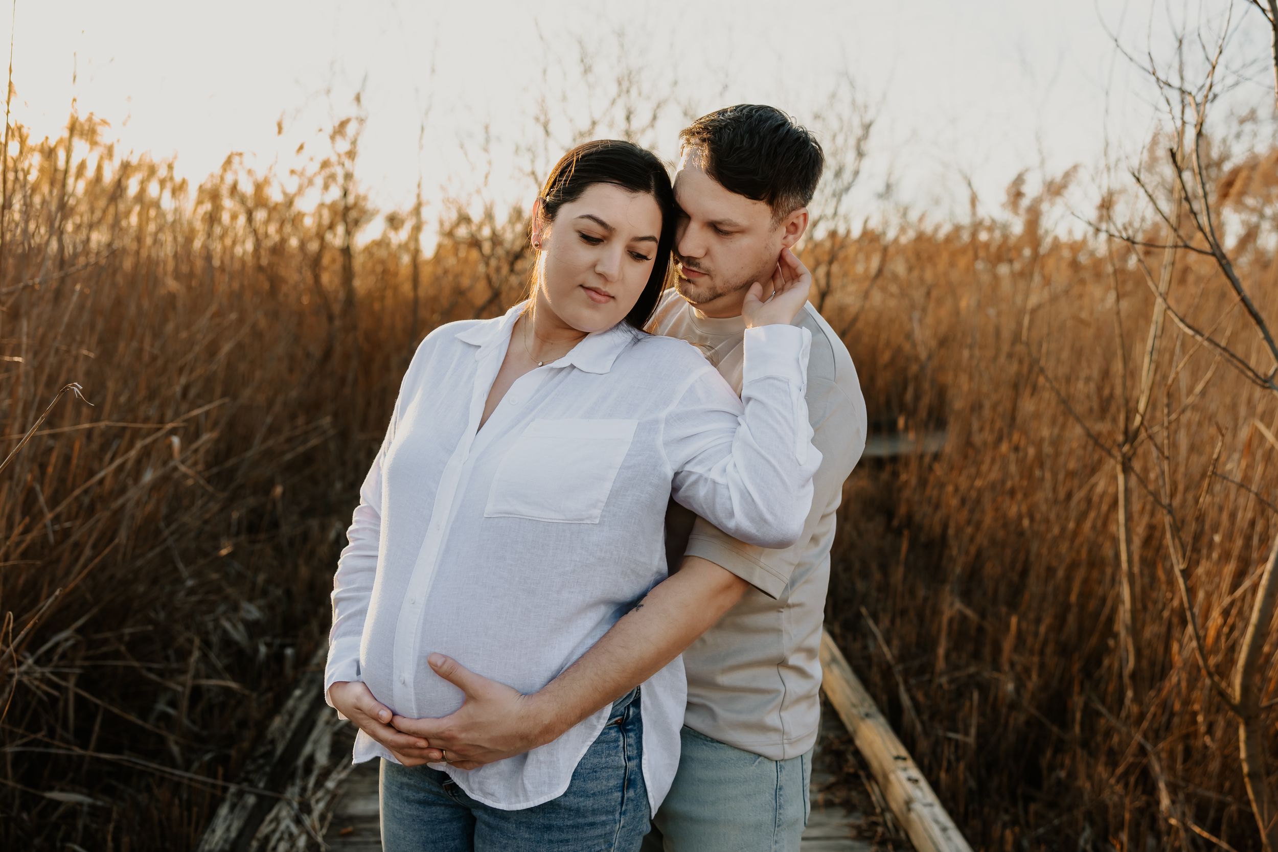 Expecting couple sharing a tender moment on a boardwalk surrounded by tall golden grass at sunset.