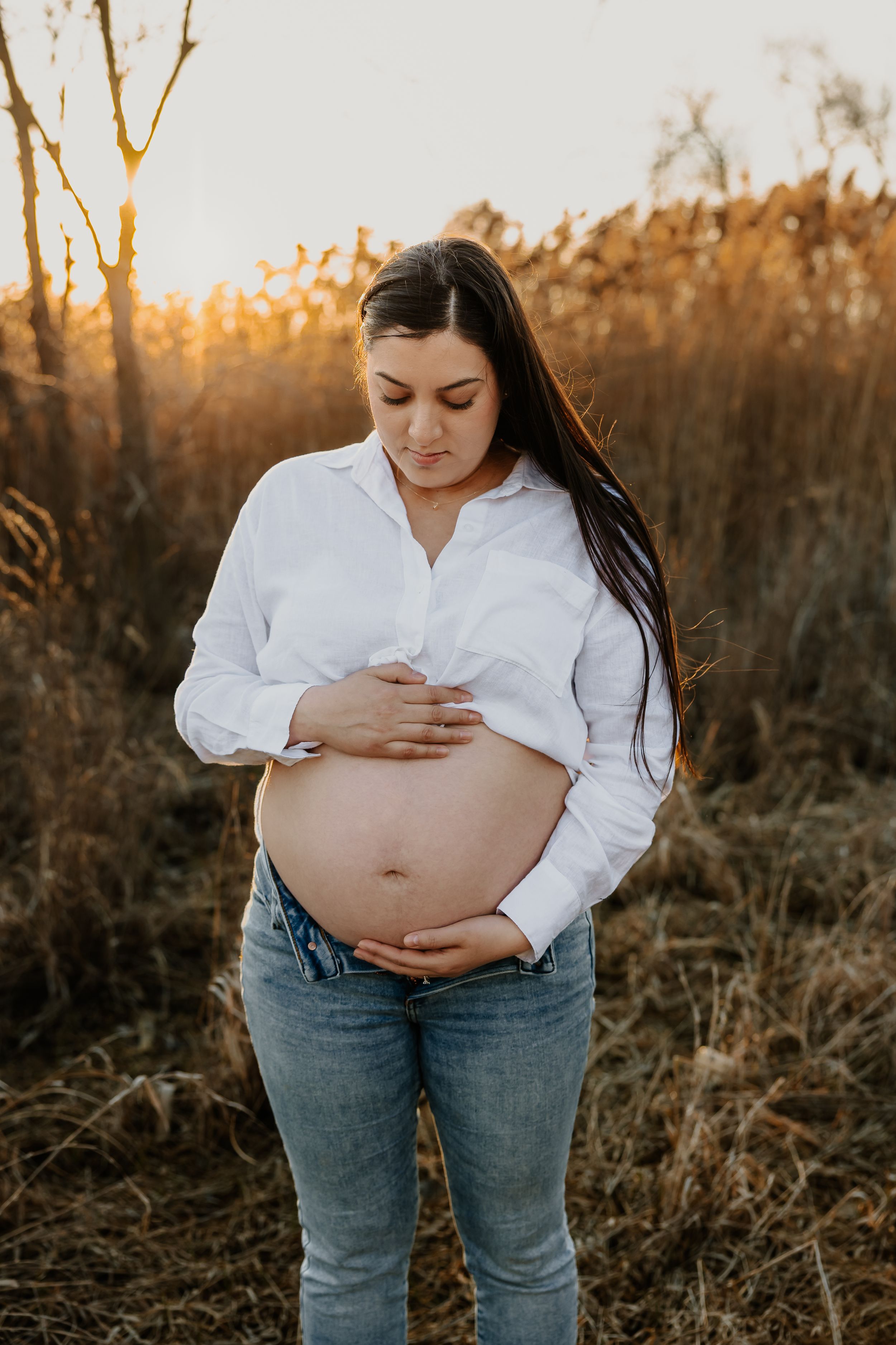 Pregnant woman in white shirt and jeans cradling her bare baby bump in a golden sunset field.