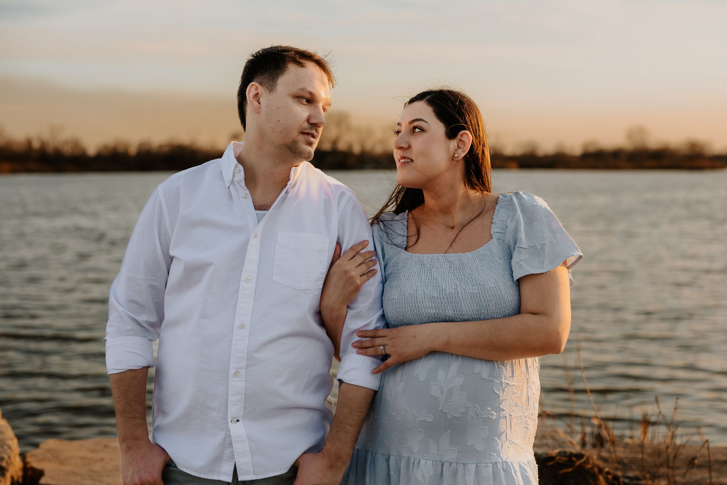 Couple sharing a tender moment by the water at sunset, woman in blue dress holding man's arm.