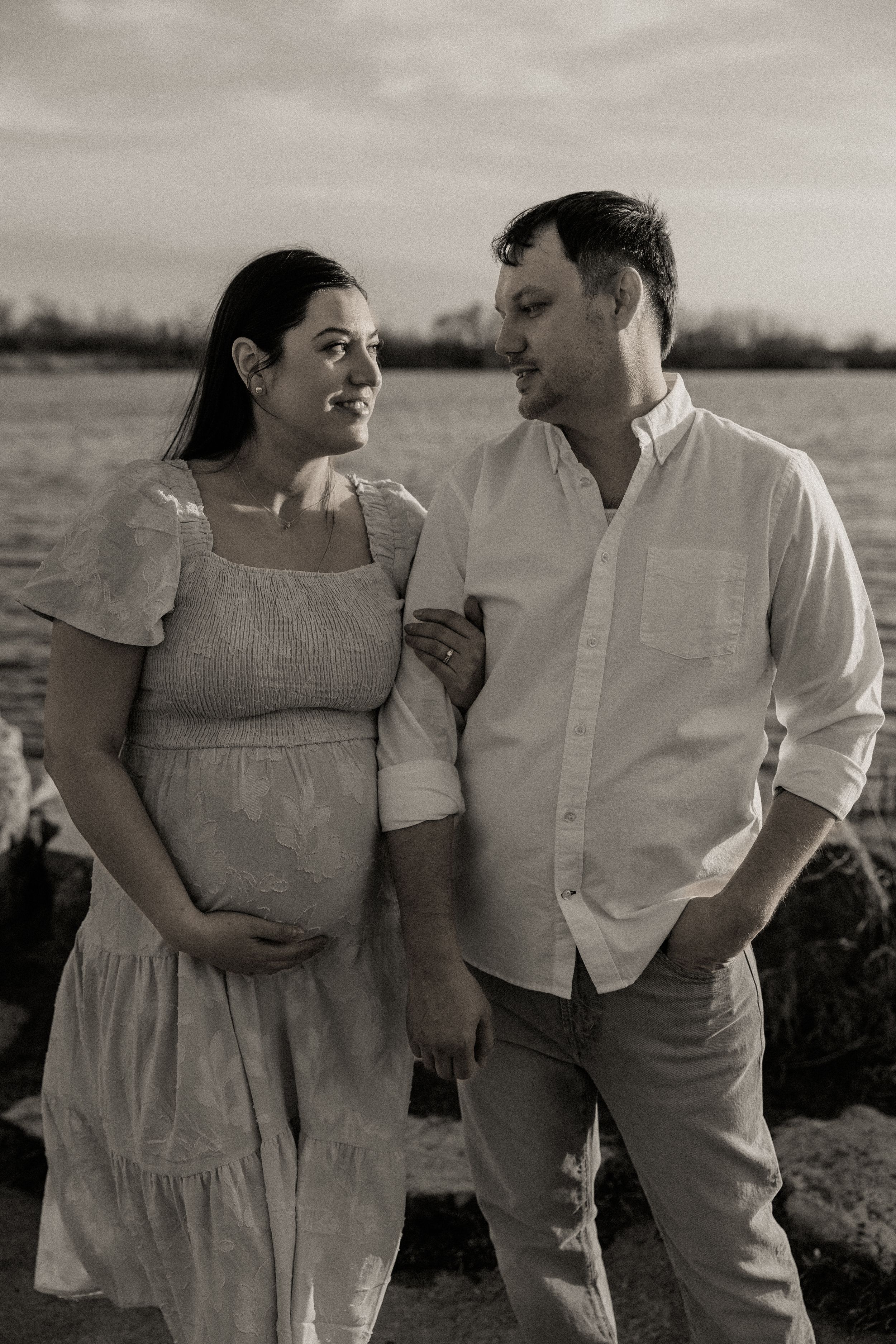 Black and white maternity photo of couple gazing at each other by the water at sunset.