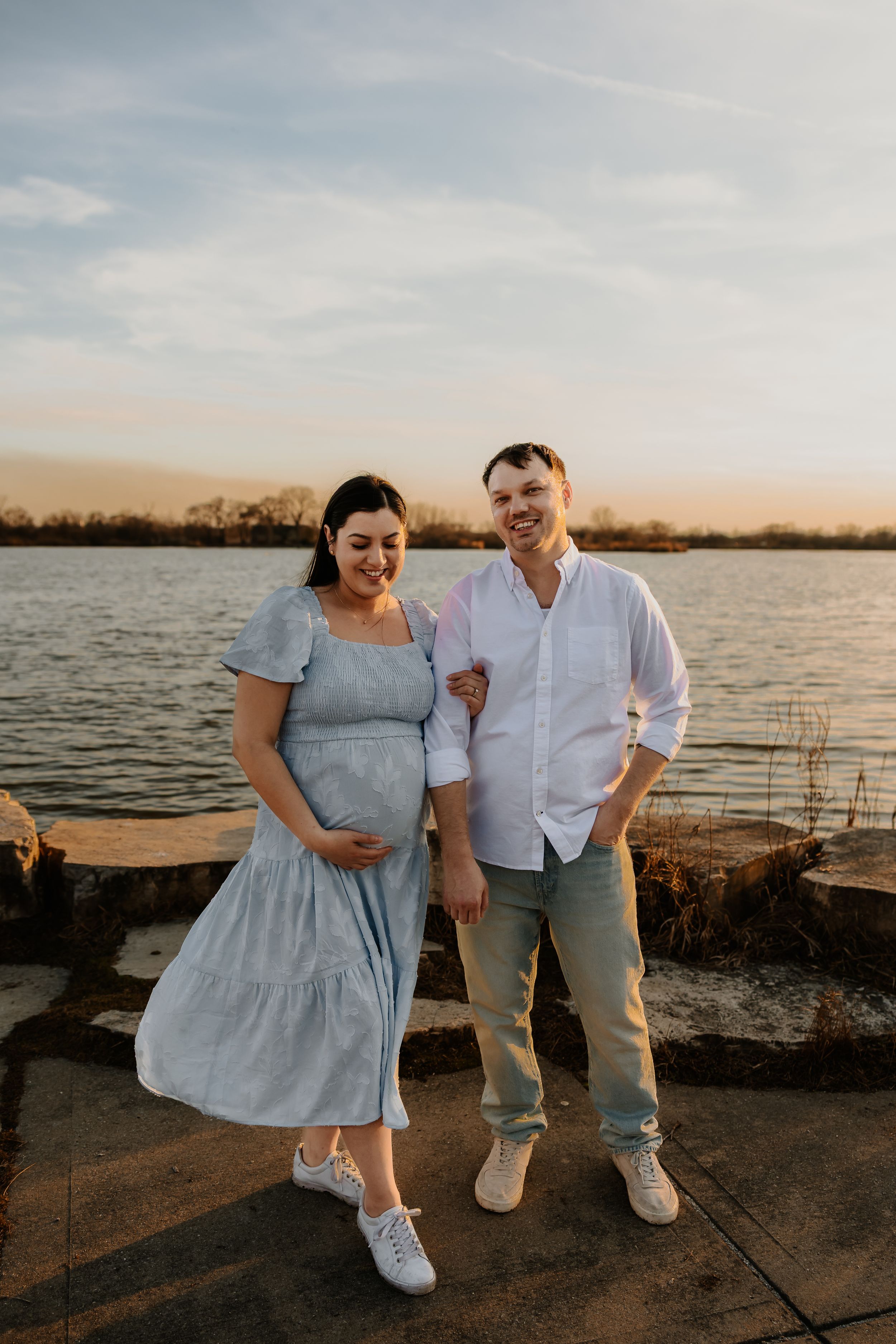 Expecting couple smiling together on waterfront at golden hour, woman in blue maternity dress.