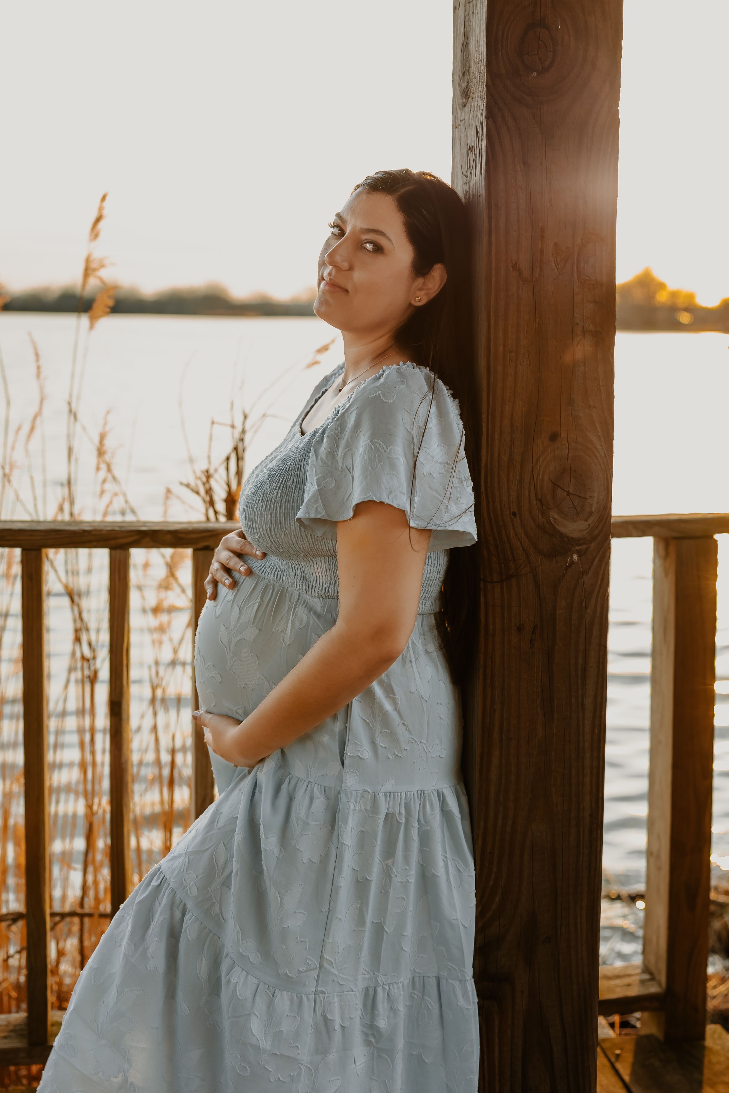 Pregnant woman in light blue dress leaning against wooden post by lake at golden hour.