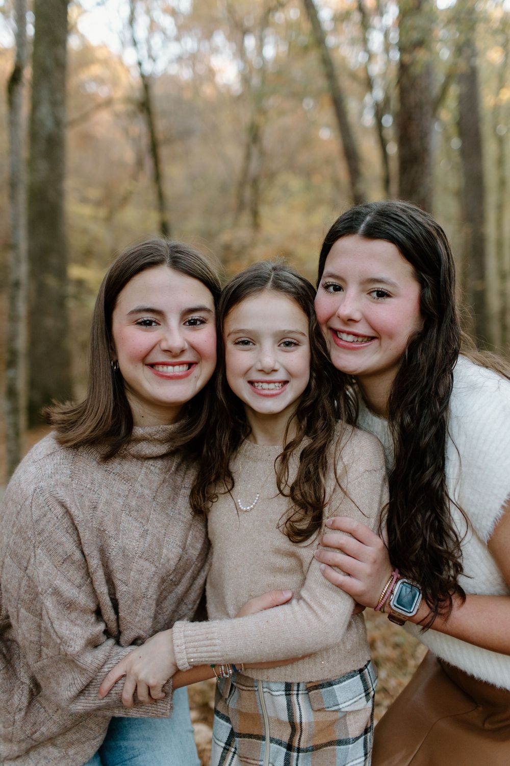 Three young girls in matching beige sweaters share hugs and smiles in wooded setting.