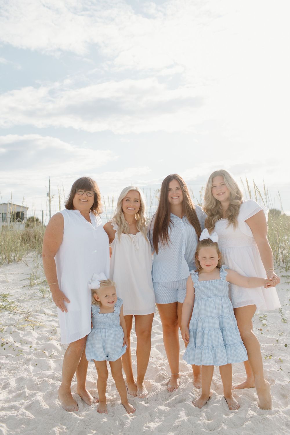 A group wearing coordinated white and light blue outfits poses together on a sunny beach with sand dunes.