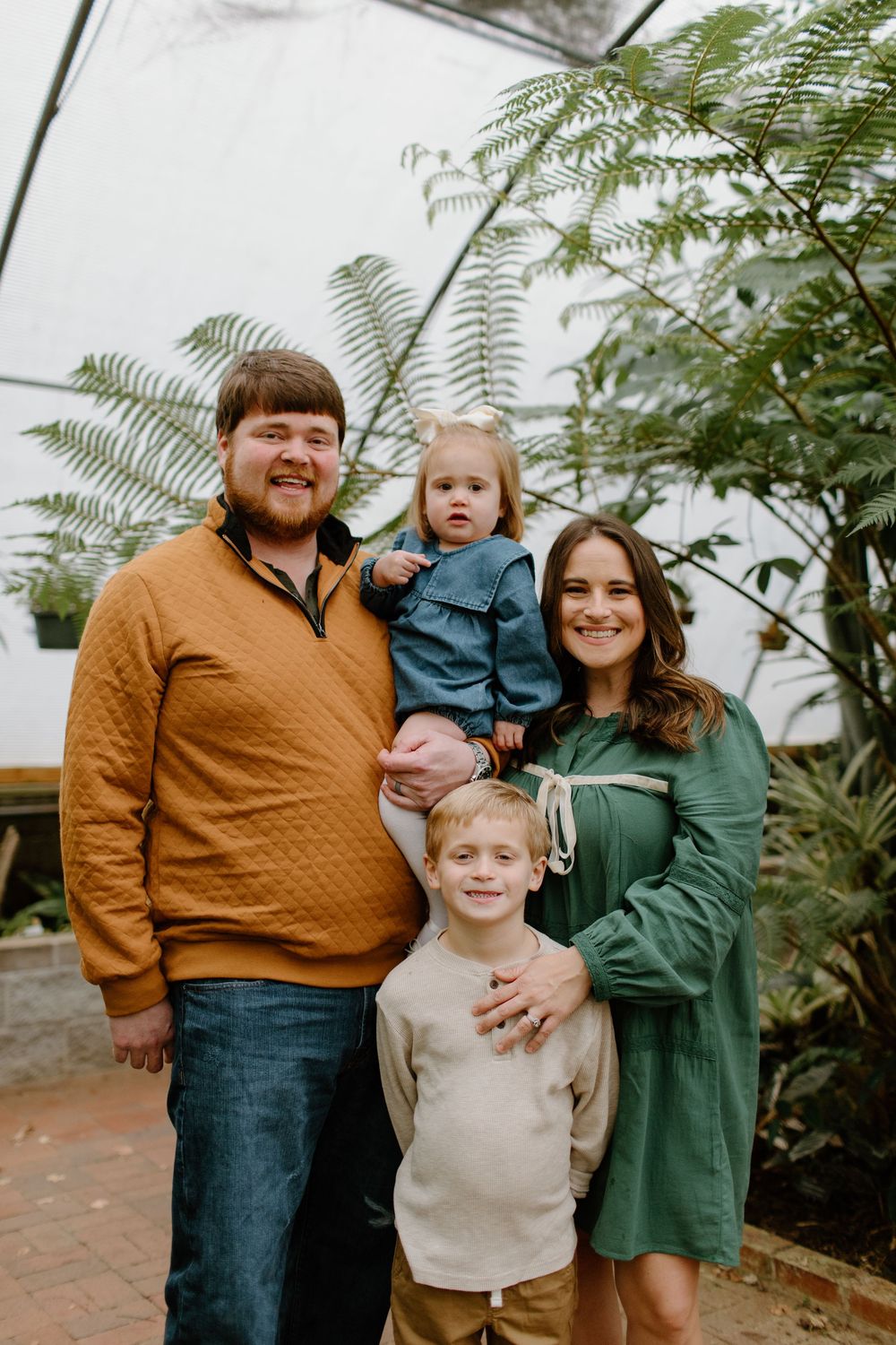 Family wearing coordinated fall colors poses together in greenhouse with plants.